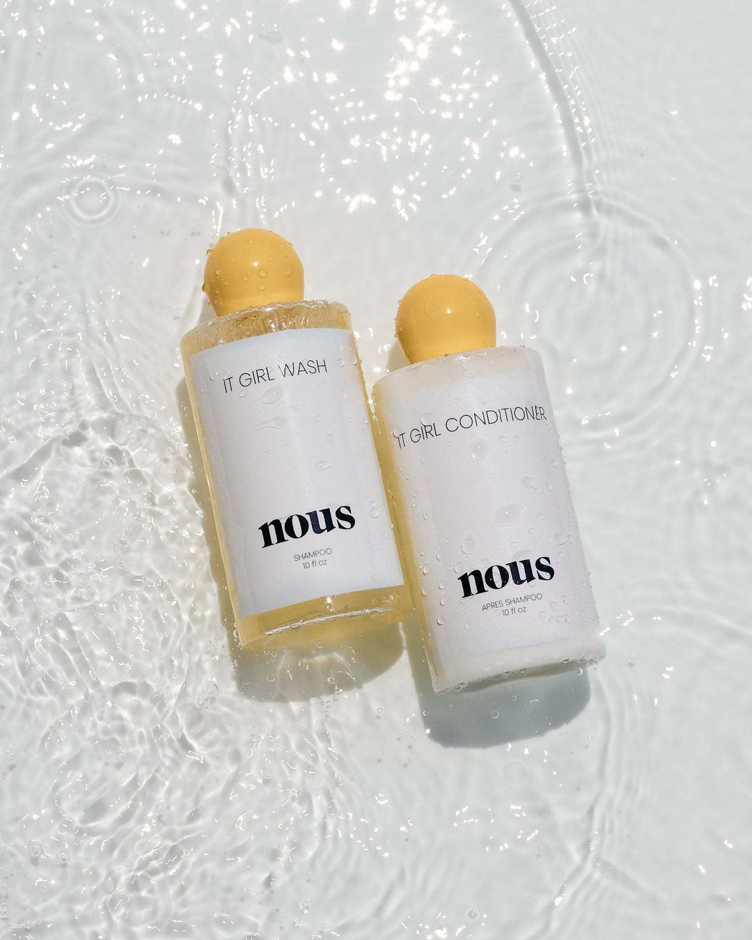 The It Girl Bundle shampoo and conditioner bottles with yellow caps laying in water, emphasizing luxury hair care. THE IT GIRL BUNDLE